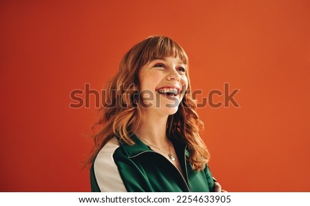 Happy young woman looking away with a smile while standing in a studio. Cheerful woman with ginger hair standing against a vibrant orange background in casual clothing.