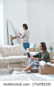 Happy Young Woman Looking At Picture In Frame While Sitting On The Floor On Background Of Her Husband Unpacking Mirror In Front Of Wall