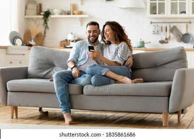 Happy young woman and man hugging, using smartphone together, sitting on cozy couch at home, smiling overjoyed wife and husband looking at phone screen, sitting on sofa in modern living room