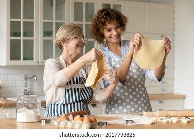 Happy young woman with mature mother cooking homemade cookies pastry in kitchen together, holding dough, excited senior mom with grown up daughter having fun, spending leisure time together