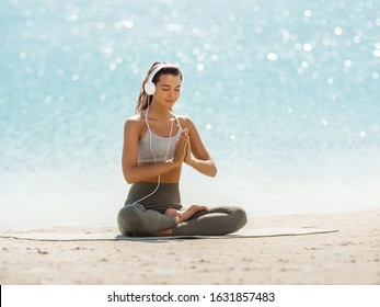 Happy Young Woman Meditating At The Seacoast. Girl Practicing Yoga On The Beach At Sunrise. Shallow DOF Copy Space Sea Background. Music For Meditation Creative Concept