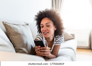 Happy Young Woman With Mobile Phone Laying On Sofa. Young Happy Woman Lie Down On Bed And Playing Smart Phone At Home. Beautiful Young Black Women Using Tablet Computer