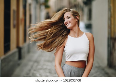 Happy young woman with moving hair in urban background. Blond girl, straight hairstyle, wearing casual clothes in the street.