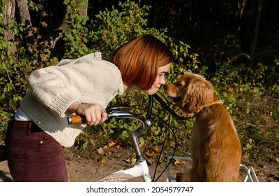 Happy young woman on the bicycle is cuddling, touching noses with her dog that sits in the basket.