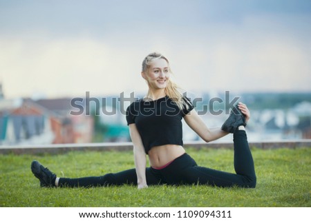 Happy young woman on the grass performs twine with the bent leg, on background cityscape