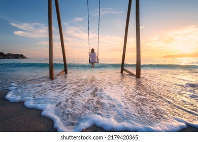 Joven feliz sobre balancín de madera en el agua, hermoso mar azul con olas, playa de arena, cielo dorado al atardecer. Vacaciones de verano en Oludeniz, Turquía. Chica montada en un columpio en la costa, agua clara. Viajes