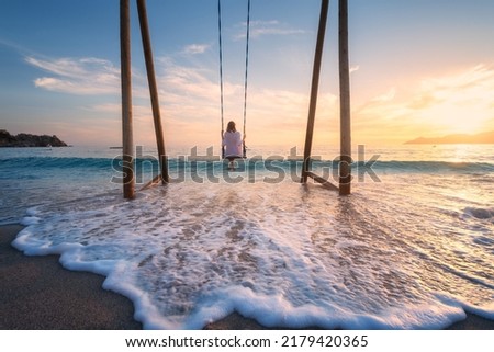 Happy young woman on wooden swing in water, beautiful blue sea with waves, sandy beach, golden sky at sunset. Summer holiday in Oludeniz, Turkey. Girl ride on a swing on sea coast, clear water. Travel