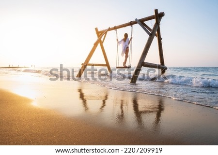 Happy young woman on wooden swing in water, beautiful blue sea with waves, sandy beach, reflection in water, golden sky at sunset. Summer in Side, Turkey. Girl ride on a swing on sea coast. Travel