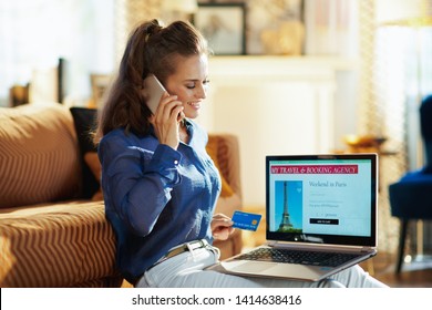 Happy Young Woman With Opened On Laptop Online Travel & Booking Agency Site Holding Credit Card Talking On A Smartphone In The Modern House In Sunny Summer Day.