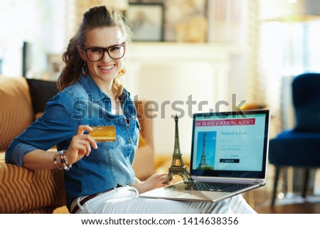 happy young woman with opened on laptop online travel & booking agency site and a souvenir of the eiffel tower holding credit card in the modern house in sunny summer day.
