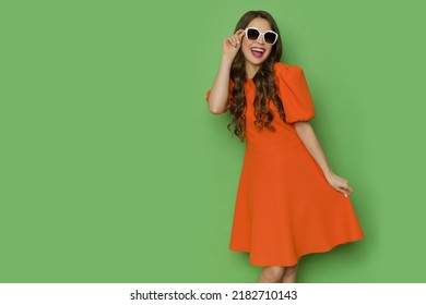 Happy young woman in orange cocktail mini dress is posing in white sunglasses and shouting. Three quarter length studio shot against green background. - Shutterstock ID 2182710143