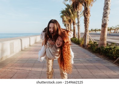 Happy young woman piggybacking her best friend on a promenade. Two cheerful girlfriends having fun together in the waterfront. Playful female friends enjoying their weekend in a coastal town.