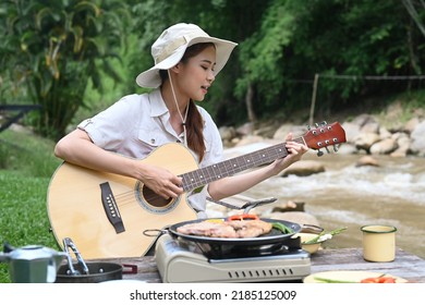 Happy Young Woman Playing Guitar During Camping Alone Near River Bank On Beautiful Day