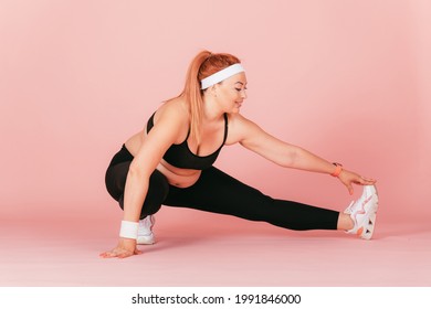 Happy Young Woman With Plus Size Body Wearing Sport Outfit Doing Leg Stretch Squat Exercise, Pink Background.