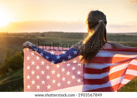 Happy young woman posing with USA national flag standing outdoors at sunset. Positive female celebrating United States independence day. International day of democracy concept.