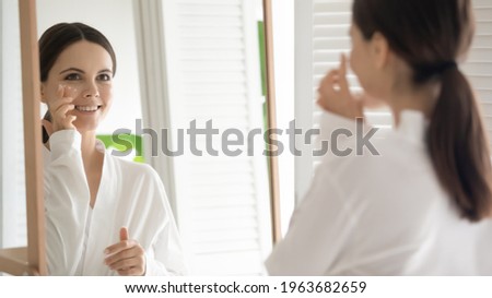 Happy young woman practicing skincare therapy procedures, applying moisturizing cosmetic lotion on face. Girl using cleaning facial cream to keep fresh healthy complexion. Mirror reflection.