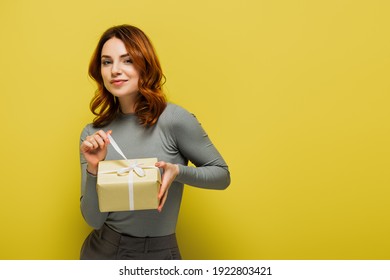 Happy Young Woman Pulling Ribbon On Present And Smiling On Yellow