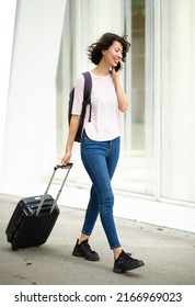 Happy Young Woman Pulling Rolling Suitcase And Carrying Backpack While Talking On Mobile Phone In City