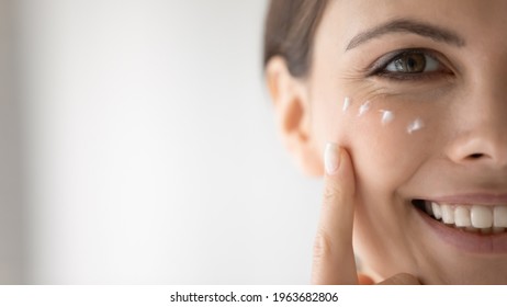 Happy young woman putting moisturizing nourishing cream, cleansing lotion on smiling face, applying cosmetics for keeping healthy fresh skin. Cosmetology, skincare concept, Close up, cropped portrait