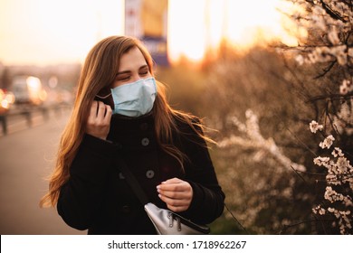 Happy young woman putting on face medical mask while standing on street in city during sunset in spring