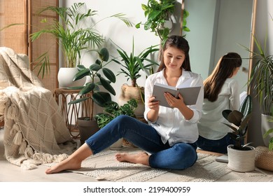 Happy Young Woman Reading Book Near Mirror And Different Houseplants On Floor In Room