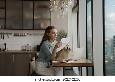 Happy young woman reading paper letter at home workplace with laptop, getting good news, bank or insurance notice. Gen Z student girl receiving approved application from college or university