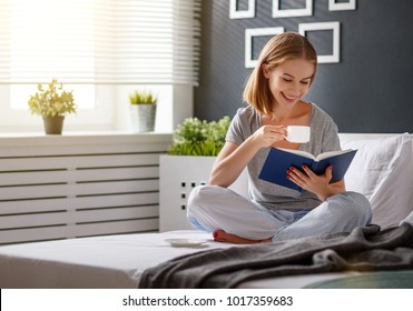 Happy young woman reads book and drinks coffee in bed in   morning