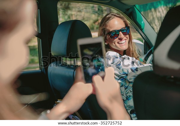 Happy young woman ready to drive car looking to\
her friend while she taking photo with a smartphone. Female\
friendship and leisure time\
concept.