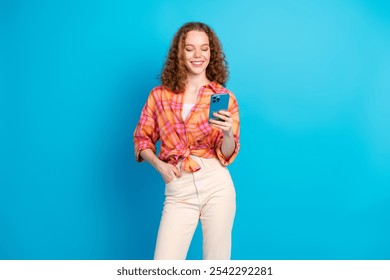 Happy young woman with red curly hair using smartphone, wearing casual plaid shirt against blue background - Powered by Shutterstock