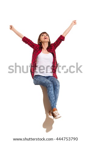 Happy young woman in red lumberjack shirt, jeans and brown sneakers sitting on a top with arms outstretched, shouting and looking up. Full length studio shot isolated on white.