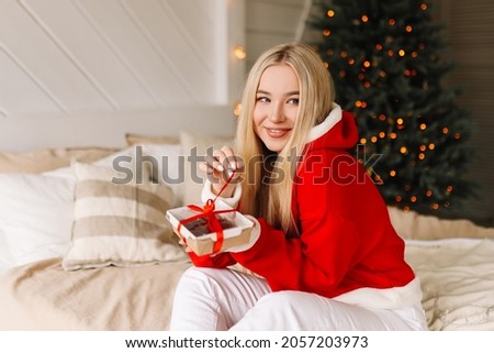 A happy young woman in a red sweater enjoys a gift box present and smiles sitting on the bed in a cozy decorated bedroom in the Christmas holidays at home. Selective focus