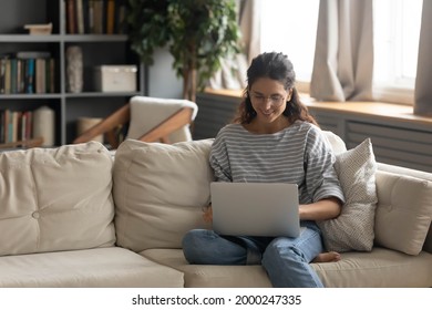 Happy Young Woman Relax On Couch At Home Browse Wireless Internet On Laptop. Smiling Millennial Female In Glasses Work Online On Computer, Talk Speak On Video Call. Technology Concept.