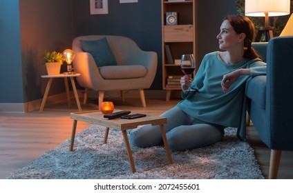 Happy Young Woman Relaxing At Home Alone, She Is Sitting On The Floor In The Living Room And Drinking A Glass Of Red Wine