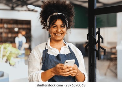 Happy young woman and restaurant owner holds her phone, expertly using mobile technology to manage her small business with ease. Female entrepreneur making success in the hospitality industry. - Powered by Shutterstock