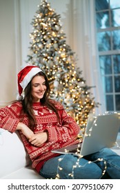 Happy Young Woman In Santa Hat On Remote Video Call On Christmas Using Laptop, Having New Year Online Virtual Party Meeting Video Call Sitting On Couch At Home.