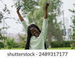 Happy young woman screams WOW holding smartphone jumping on grass in city park. African American with dreadlocks expresses astonishment due to good news. Lady makes energetic moves experiencing joy