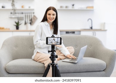 Happy young woman sharing her success with subscribers online. Positive caucasian female influencer recording video on modern smartphone while sitting on couch with laptop. - Powered by Shutterstock