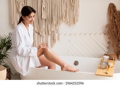 Happy Young Woman Shaving Legs Removing Hair With Razor Sitting On Bathtub In Modern Bathroom At Home. Hygiene, Pampering And Beauty Routine. Aftershave Skincare Concept. Side View