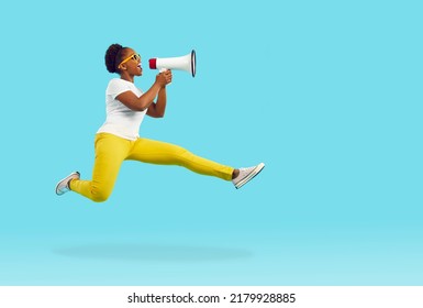 Happy Young Woman Shouts Through Megaphone. Funny Crazy Afro Lady In T Shirt, Yellow Pants And Sunglasses Jumps High In Air And Yells In Loudspeaker Isolated On Bright Blue Color Background