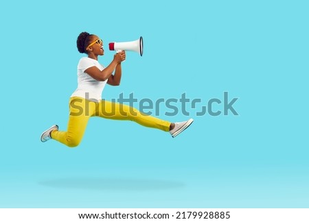 Happy young woman shouts through megaphone. Funny crazy Afro lady in T shirt, yellow pants and sunglasses jumps high in air and yells in loudspeaker isolated on bright blue color background