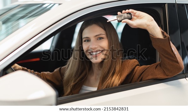 Happy young woman showing car key from auto\
window, purchasing new vehicle at showroom store, banner. Cheery\
millennial female client sitting in drivers seat, buying luxury\
automobile at dealership