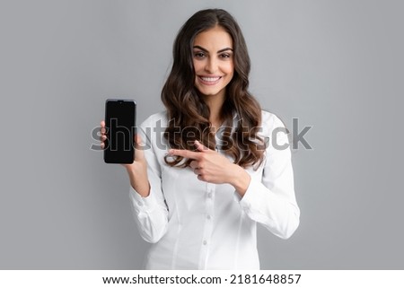 Happy young woman showing empty smartphone screen. Smiling young brunette business woman girl in shirt isolated on gray background, studio portait. Beauty girl using mobile phone, typing sms message.