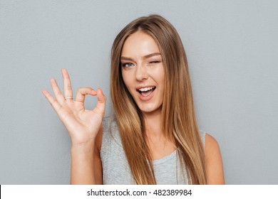 Happy Young Woman Showing Ok Sign With Fingers An Winking Isolated On A Gray Background