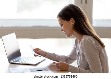 Happy Young Woman Sit At Table Working On Laptop Reading Good News In Paper Letter, Smiling Girl Receive Pleasant News Get Mail From University Or College, Excited Female Look Through Great Job Offer