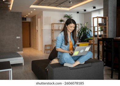 Mujer joven feliz sentada con las piernas cruzadas en un sofá acogedor, trabajando desde casa con su computadora portátil en un apartamento de diseño elegante, disfrutando de un estilo de vida cómodo y productivo