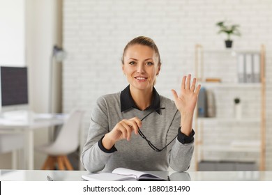 Happy Young Woman Sitting At Desk, Waving Hand At Camera. School Teacher, Online Tutor, University Professor, College Student, Businesswoman, Vlogger Or Remote Worker Says Hello In Video Call Meeting