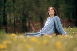 
Happy Young Woman Sitting In The Grass Enjoining Nature. Carefree Person Resting In A Countryside Field Experiencing Mindfulness And Calmness 
