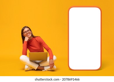 Happy young woman sitting with laptop and looking at blank screen of huge phone, mockup for app, isolated on yellow background
