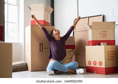 Happy Young Woman Sitting In New Apartment And Raising Arms In Joy After Moving In. Joyful And Excited African Girl Moving To New Home. Black Woman Sitting On Floor In Her House.