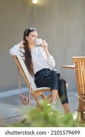 Happy Young Woman Sitting On The Terrace Of The Cafe In Sunny Day And Drinking Coffee. Woman Enjoying First Spring Sun Outdoor. 